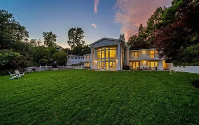 Large modern house with lit windows at dusk, set behind a wide green lawn and trees