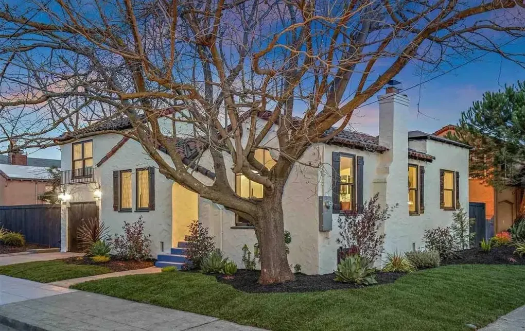 White house with lit windows, large bare tree, and sidewalk in front at dusk