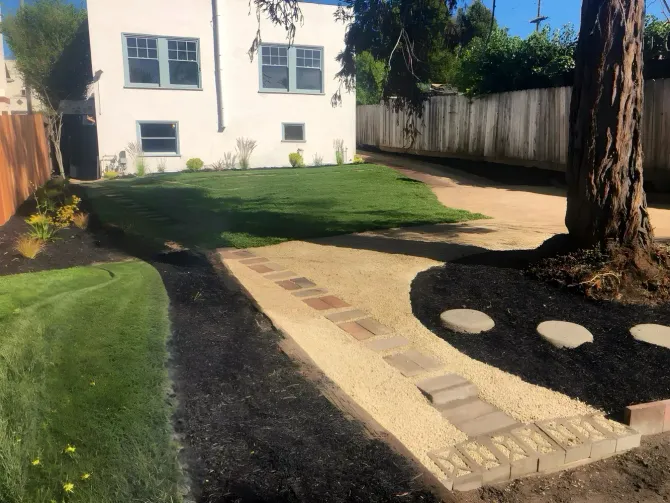 Backyard path and lawn beside a white house, with a tree and wood fence