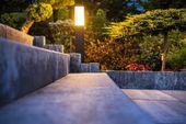 Lit stone walkway beside a glowing garden lamp and low wall at dusk