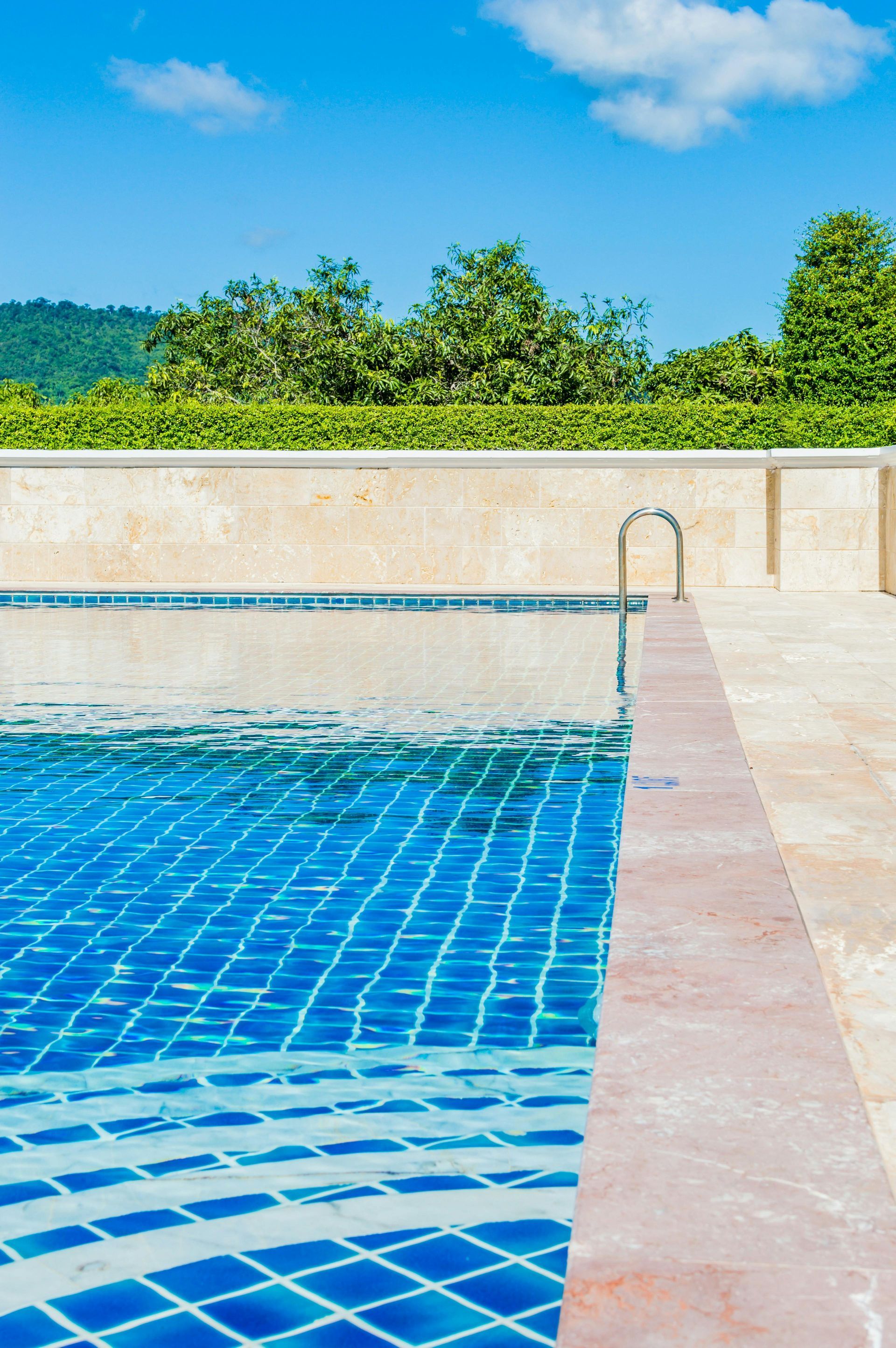 A blue-tiled swimming pool, clear sky, beige wall, and green foliage.