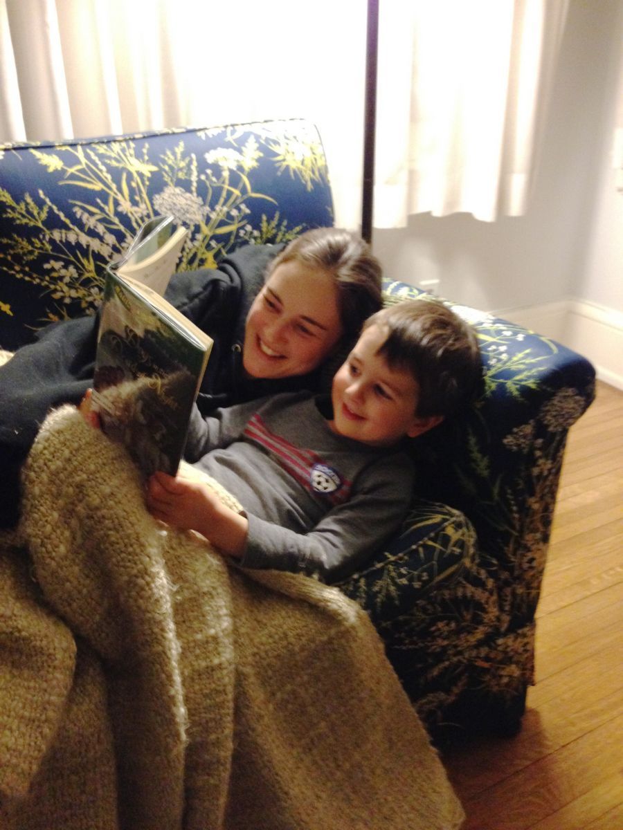 Two children under a blanket on a couch, reading a book by a sunlit window.
