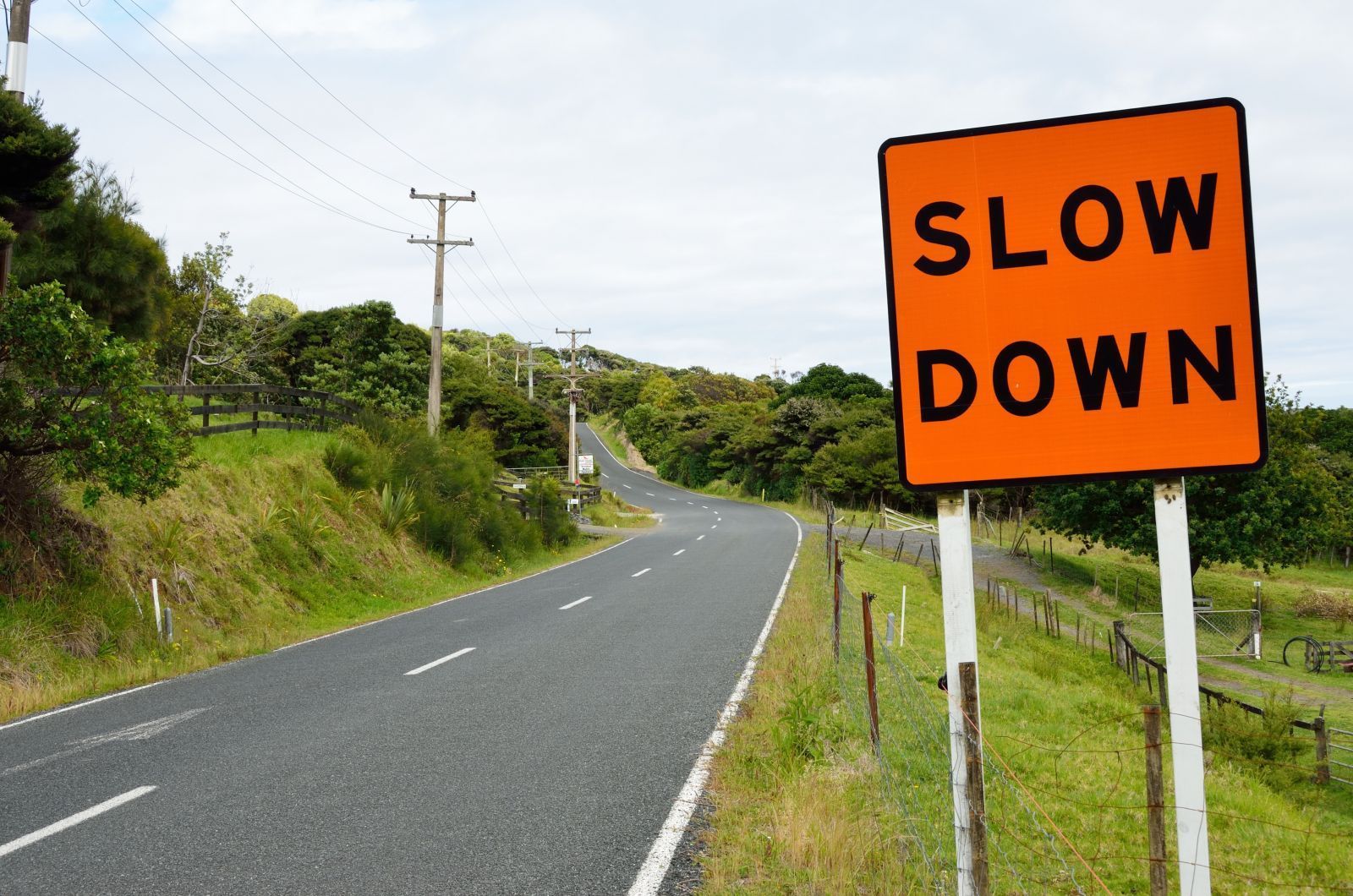 Curving rural road with orange “SLOW DOWN” sign on the right and green hillsides nearby