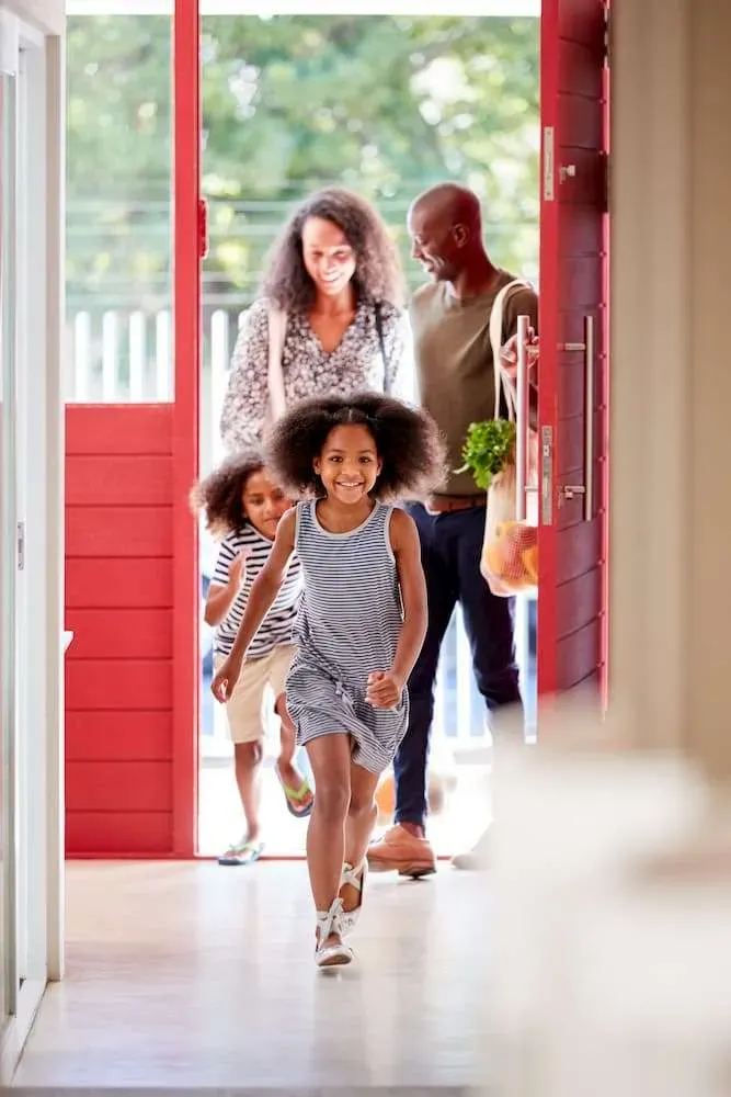 Family entering a home through a red door. Two children run ahead, while parents follow carrying groceries.