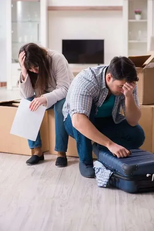Couple overwhelmed, heads in hands, surrounded by moving boxes and luggage.