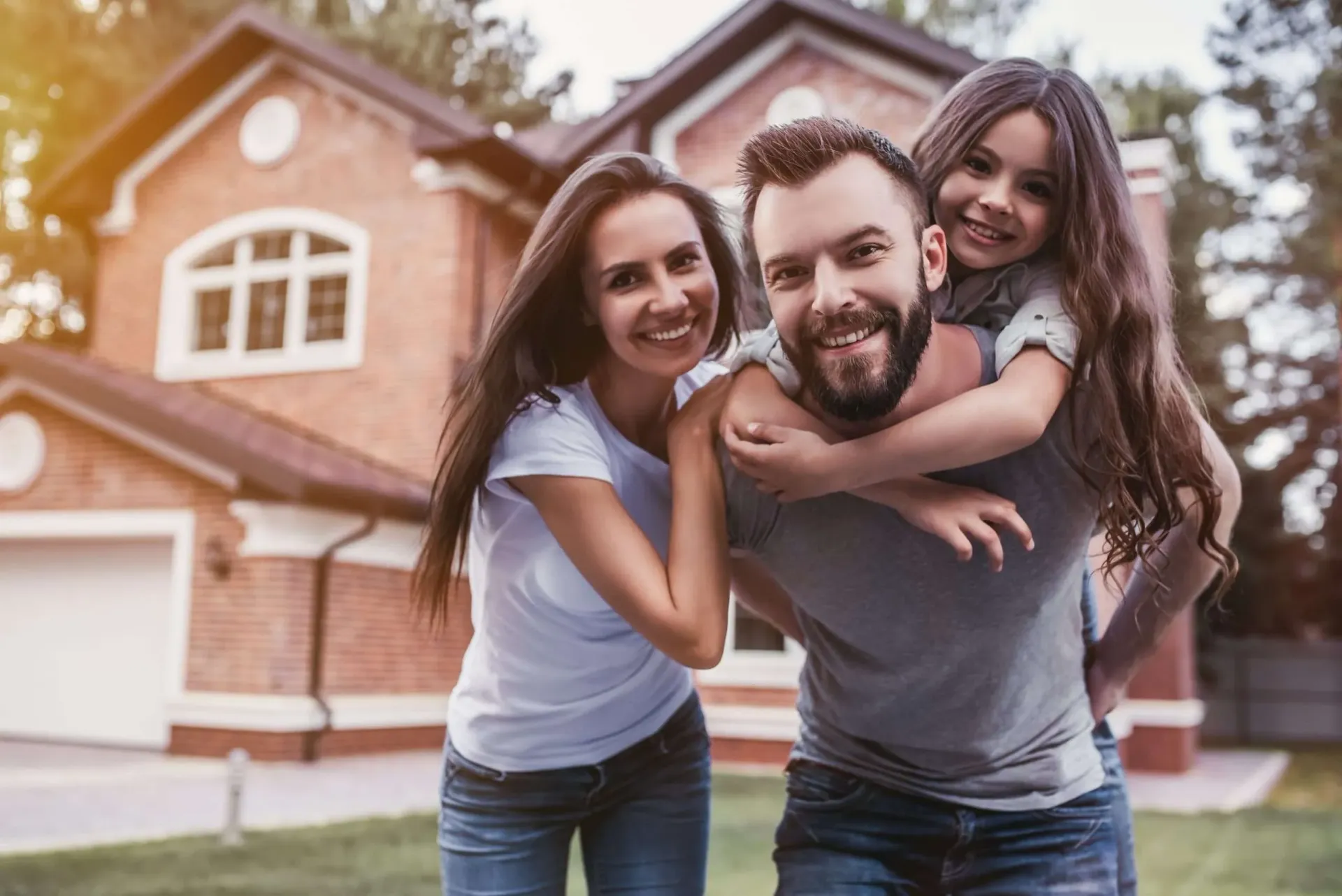Family of three smiling in front of a brick house; the father gives the daughter a piggyback ride.