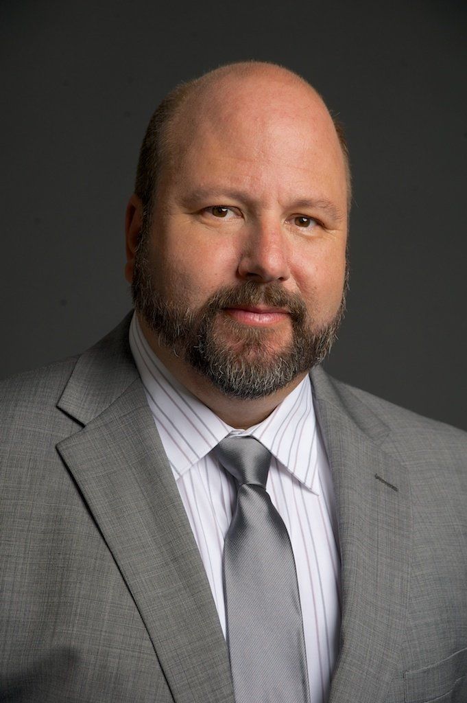 Man in gray suit and tie, dark beard, looking at the camera.