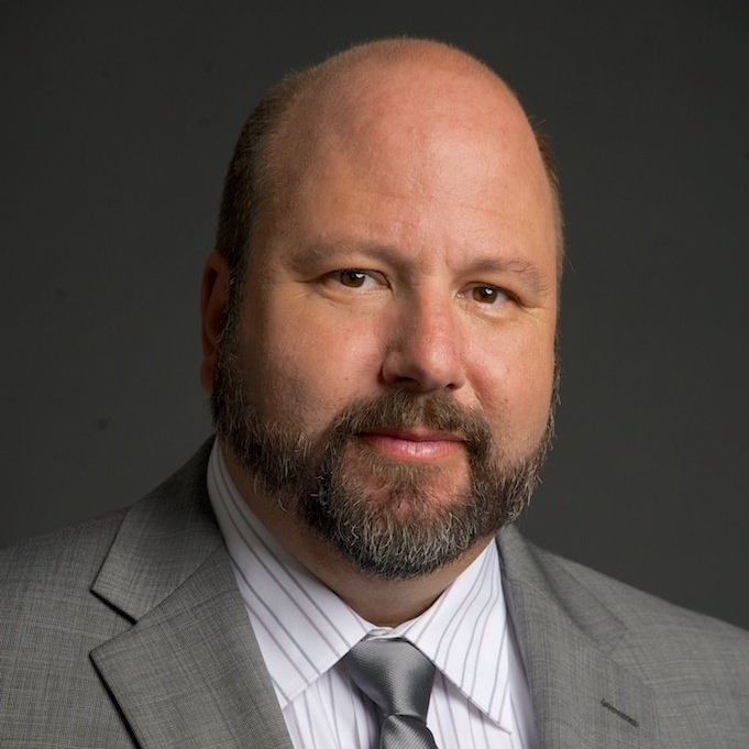 Man with a beard in a gray suit and tie, looking at the camera against a gray backdrop.