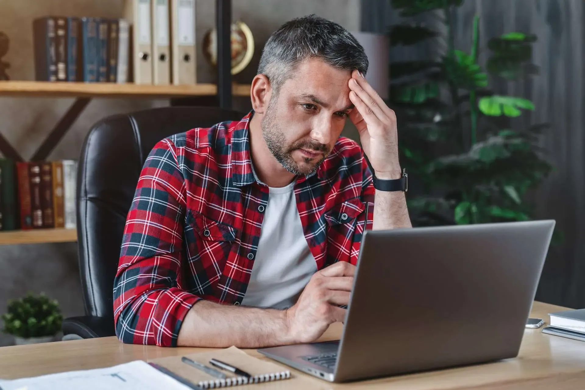 Man in red plaid shirt looking stressed at a laptop in a home office.