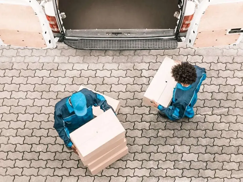 Two movers loading cardboard boxes into a van parked on a brick patio.