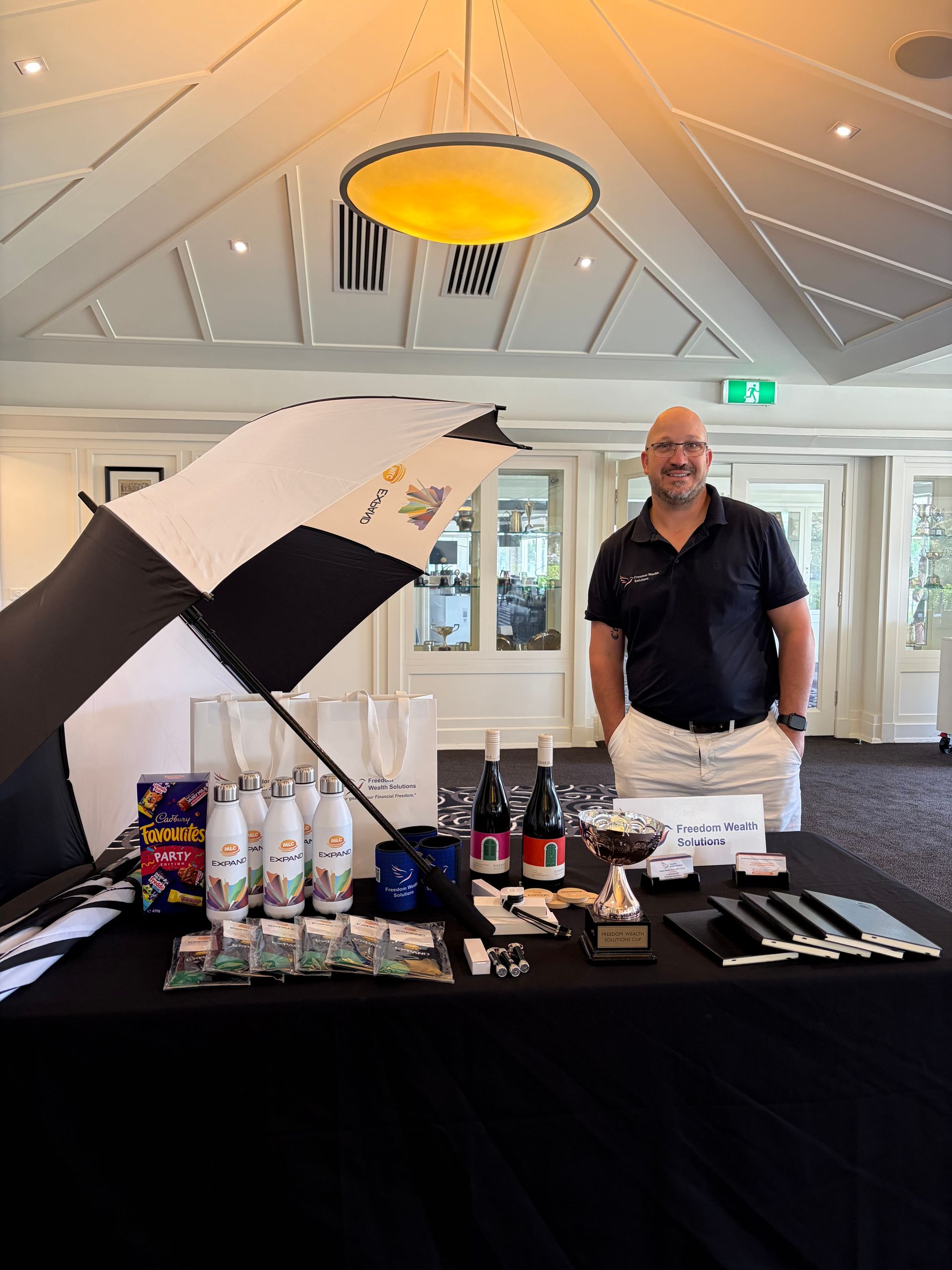 A person stands behind a table displaying event prizes, including branded water bottles, wine, and a trophy, indoors. — Freedom Wealth Solutions in Mornington, VIC