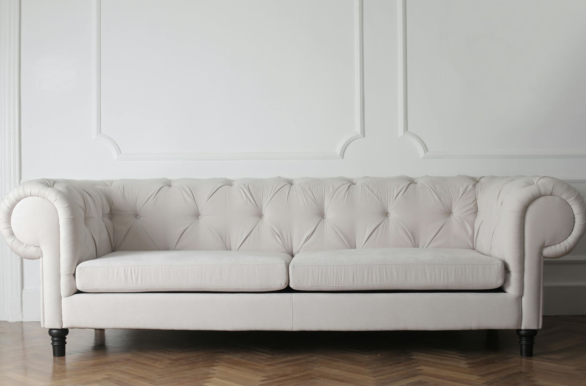 A white tufted chesterfield sofa stands against a plain white wall on a wooden herringbone floor.
