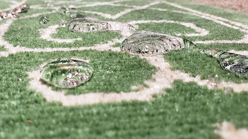 Close-up of dew droplets on grass, showing a small ground-level scene with a spiral pattern in the background