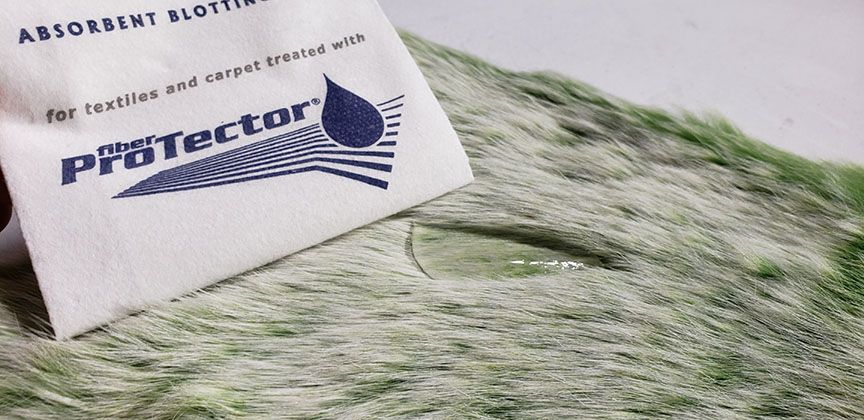 White absorbent blotter on green carpet, labeled “Protector” with black text and blue logo.