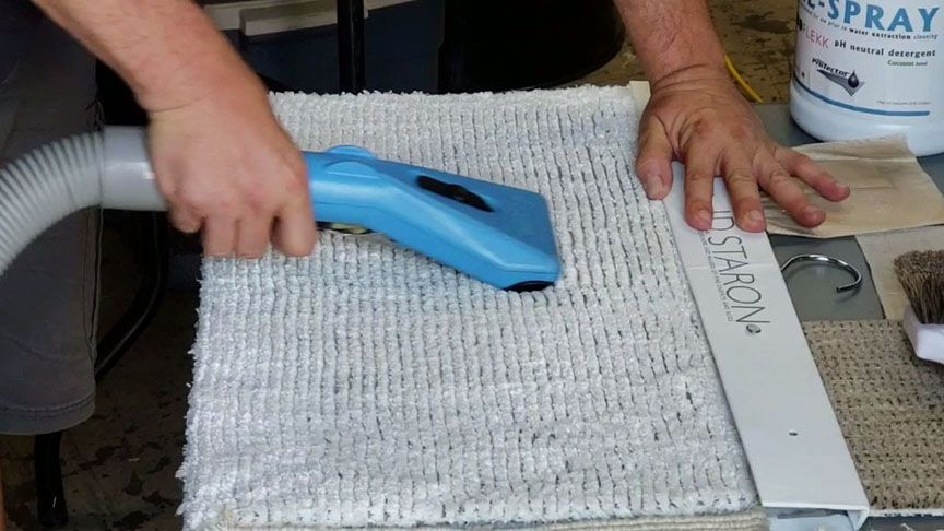 Hands vacuuming a gray carpet with a blue upholstery nozzle beside a spray bottle and metal ruler.