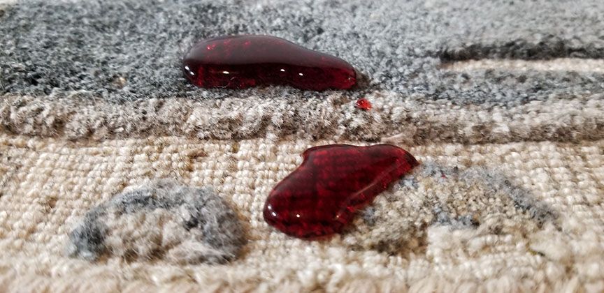 Two dark red drops of liquid on a beige carpet near a gray rug edge