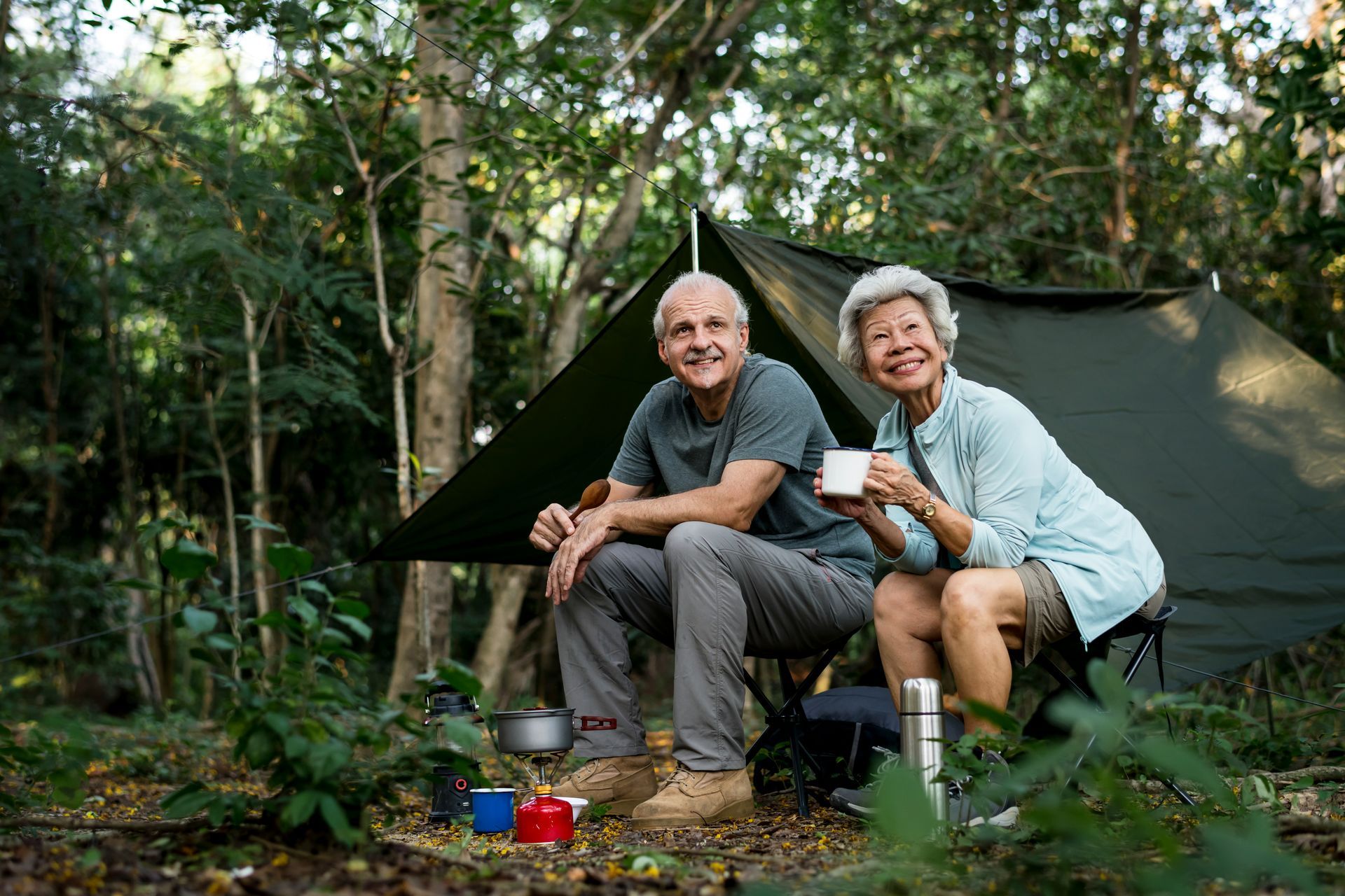 Couple camping in a forest: smiling, sitting by a tent, holding mugs.