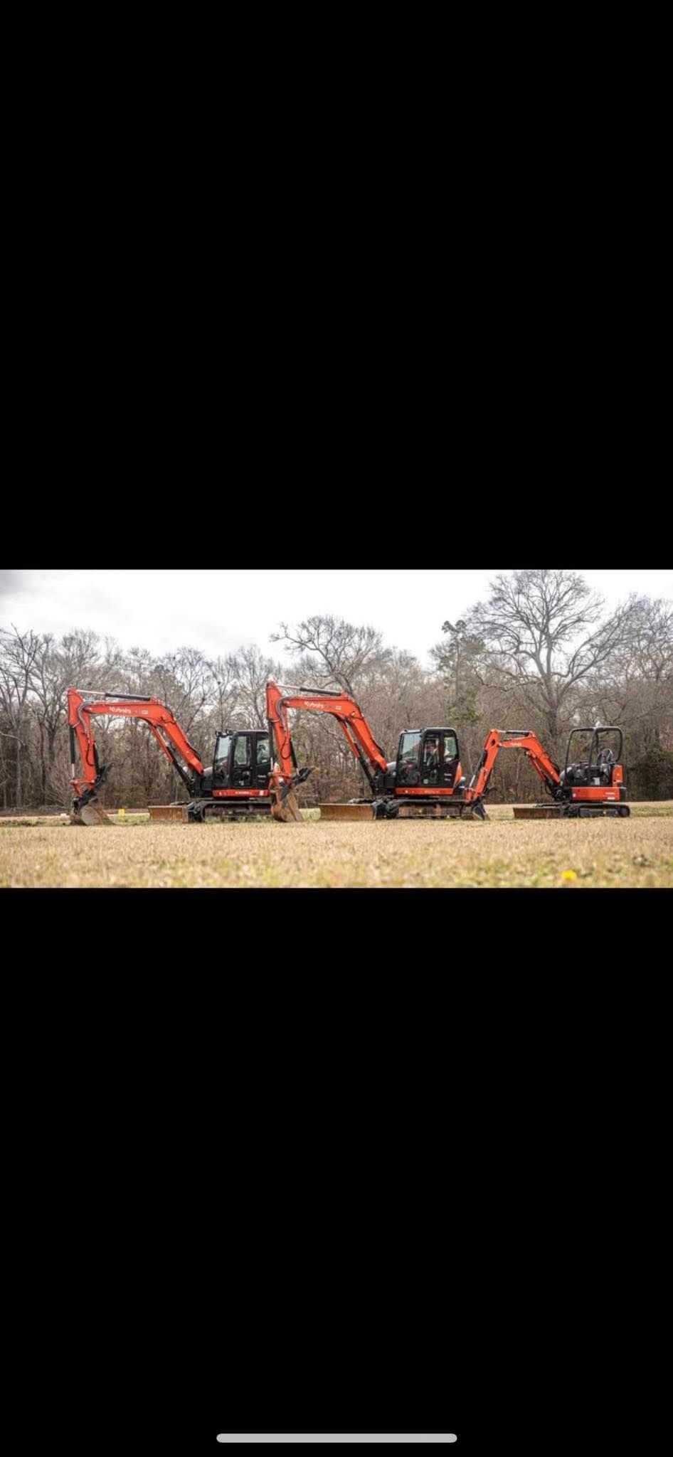 Three orange Hitachi excavators of varying sizes parked in a line on a dry, grassy field against a backdrop of trees.