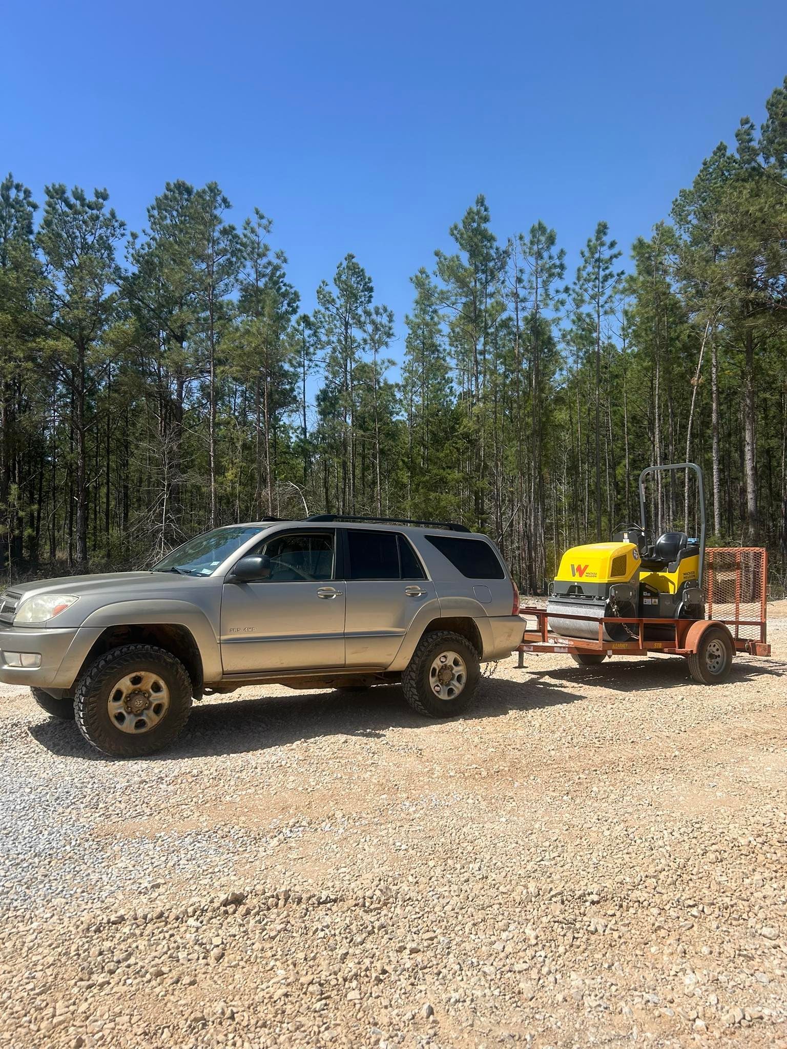 A gold SUV parked on a gravel lot towing a small trailer carrying yellow industrial machinery against a forest backdrop.
