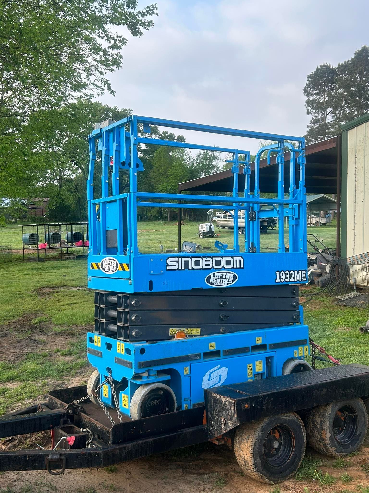 A blue Sinoboom electric scissor lift loaded on a black flatbed utility trailer in a grassy field.