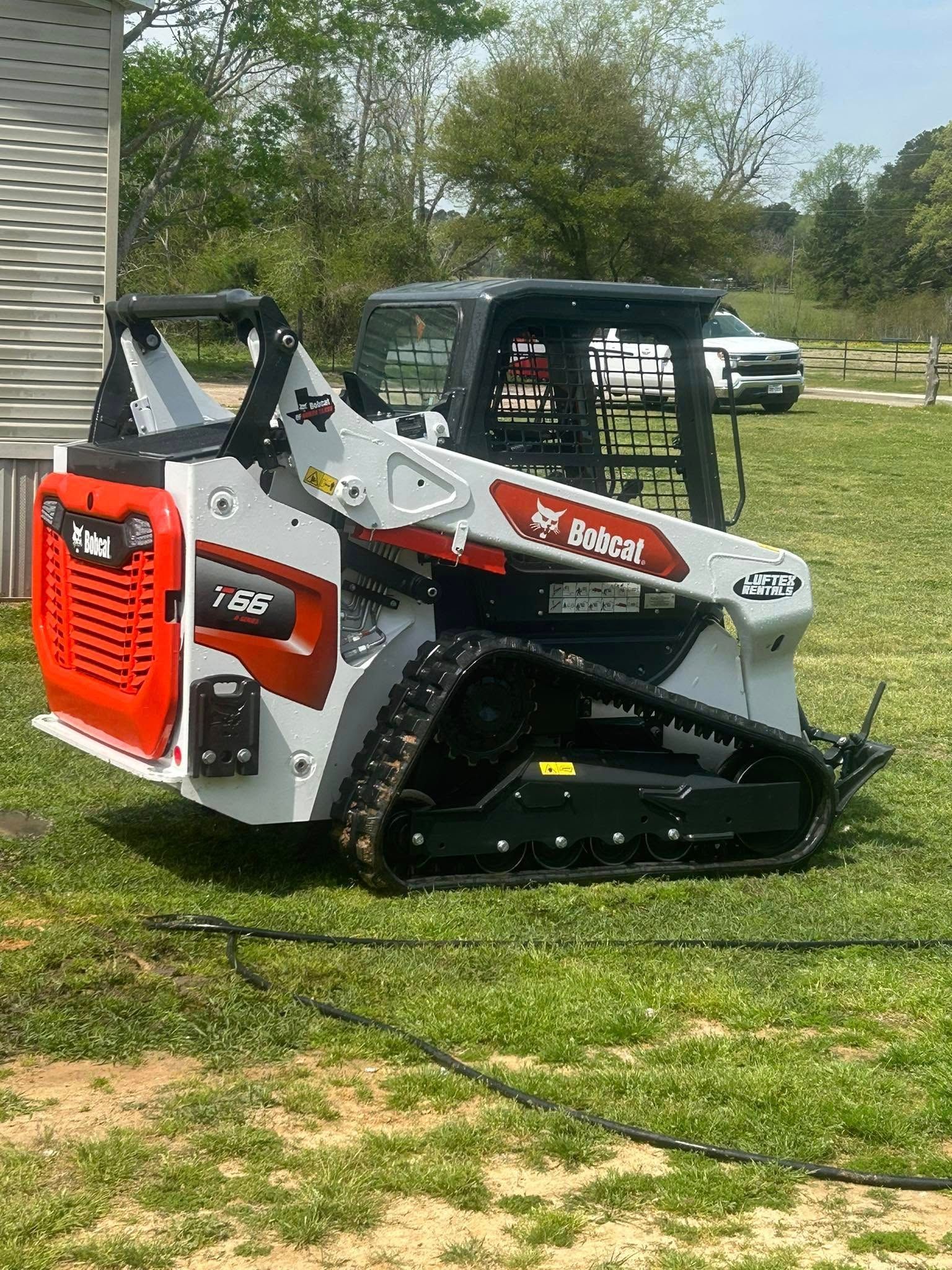 A white and red Bobcat compact track loader parked on a grassy field with trees and a white vehicle in the background.