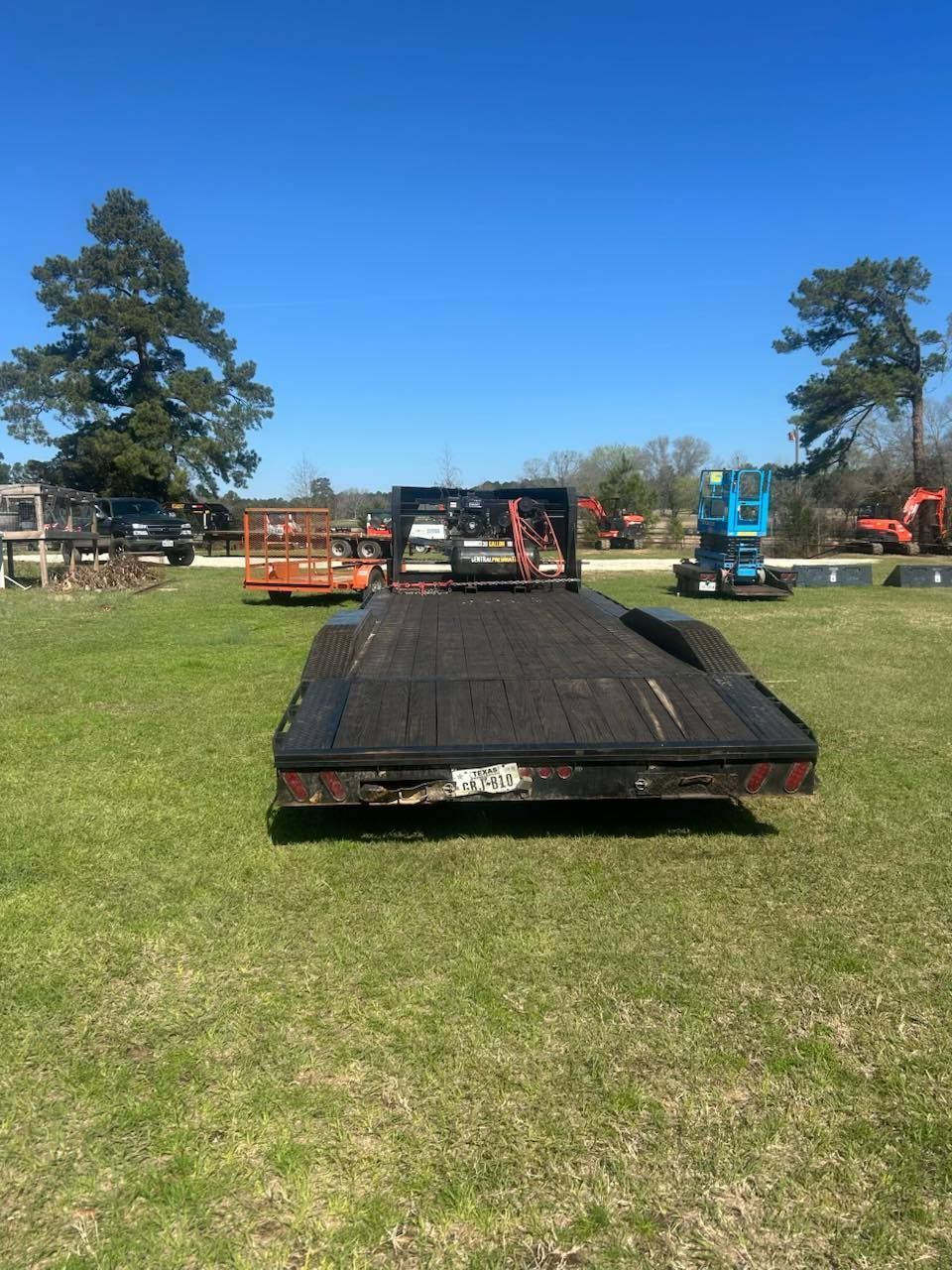 A flatbed trailer sits on a grass lot under a clear blue sky, with construction equipment visible in the background.