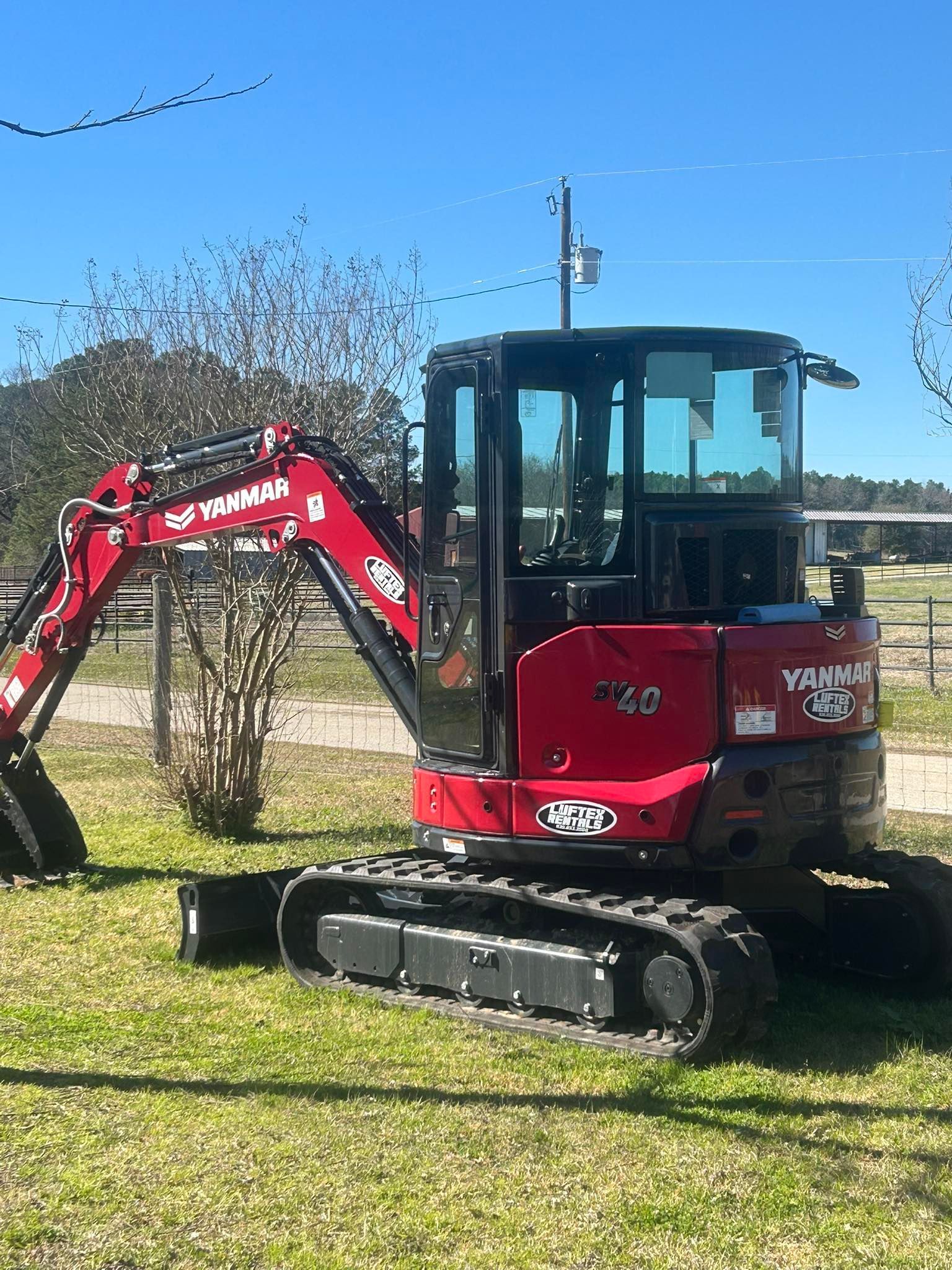 A red Yanmar excavator sits on a grassy field under a bright blue sky.
