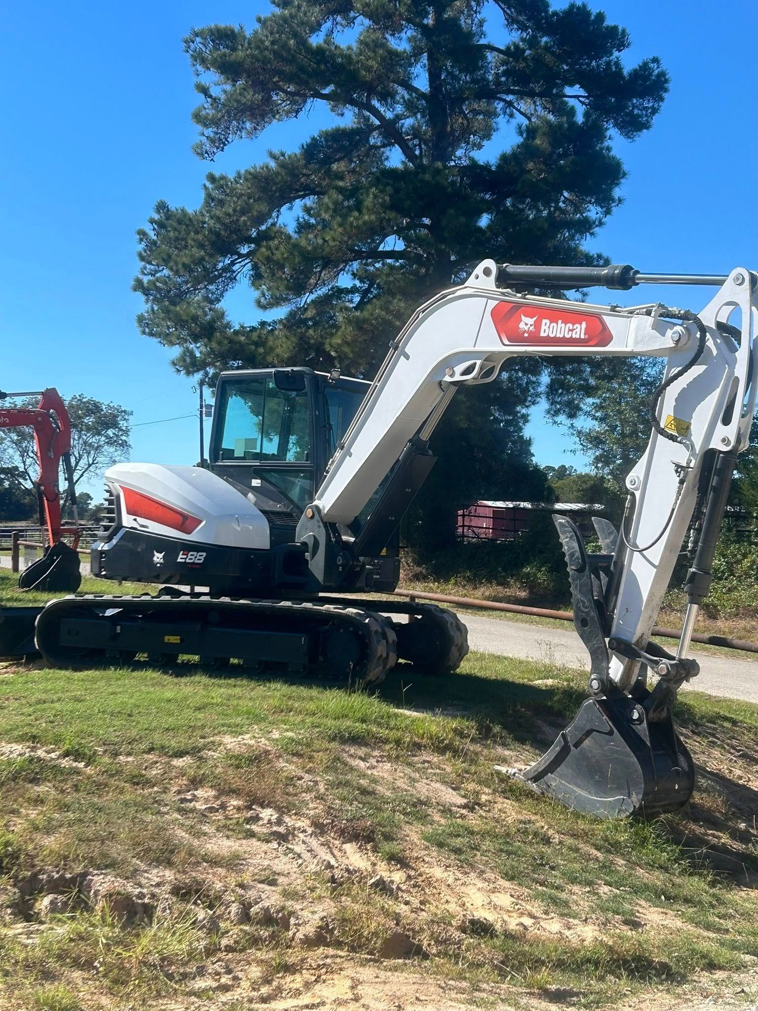 A white Bobcat excavator parked on a grassy dirt field under a clear blue sky, with a tall tree in the background.