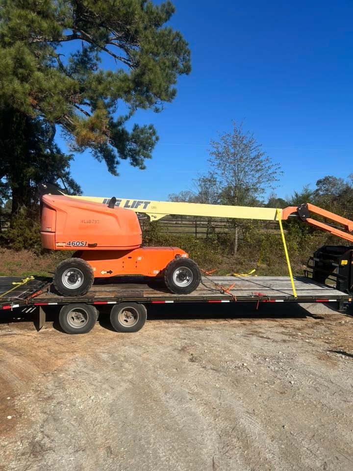 An orange JLG boom lift secured with yellow straps on a flatbed trailer against a clear blue sky.