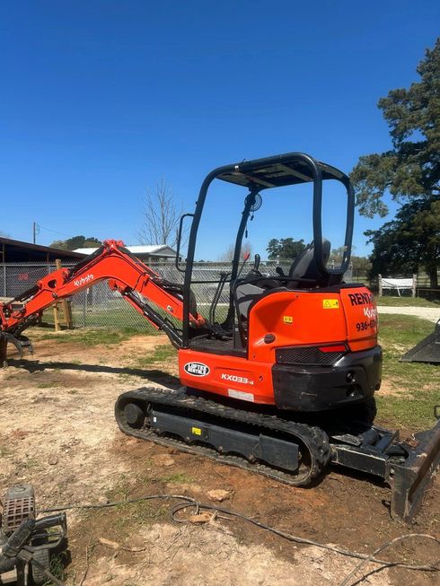 A bright orange mini excavator with black tracks parked on dirt in a rural, sunny outdoor setting.