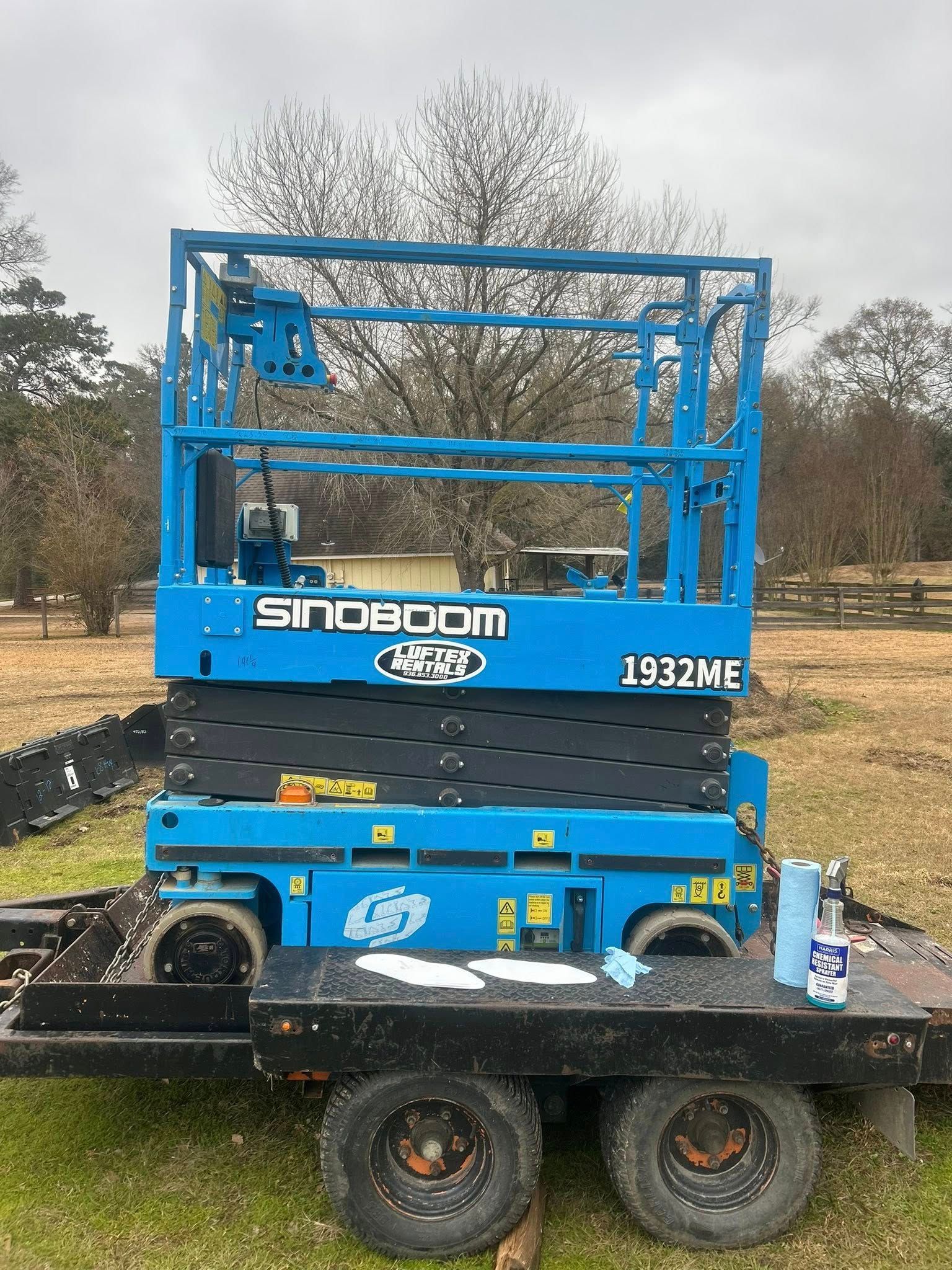 A blue Sinoboom 1932ME scissor lift loaded on a black utility trailer in a grassy field.