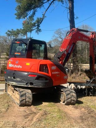 An orange Kubota KX080 mini excavator parked on a dirt lot with a clear blue sky in the background.