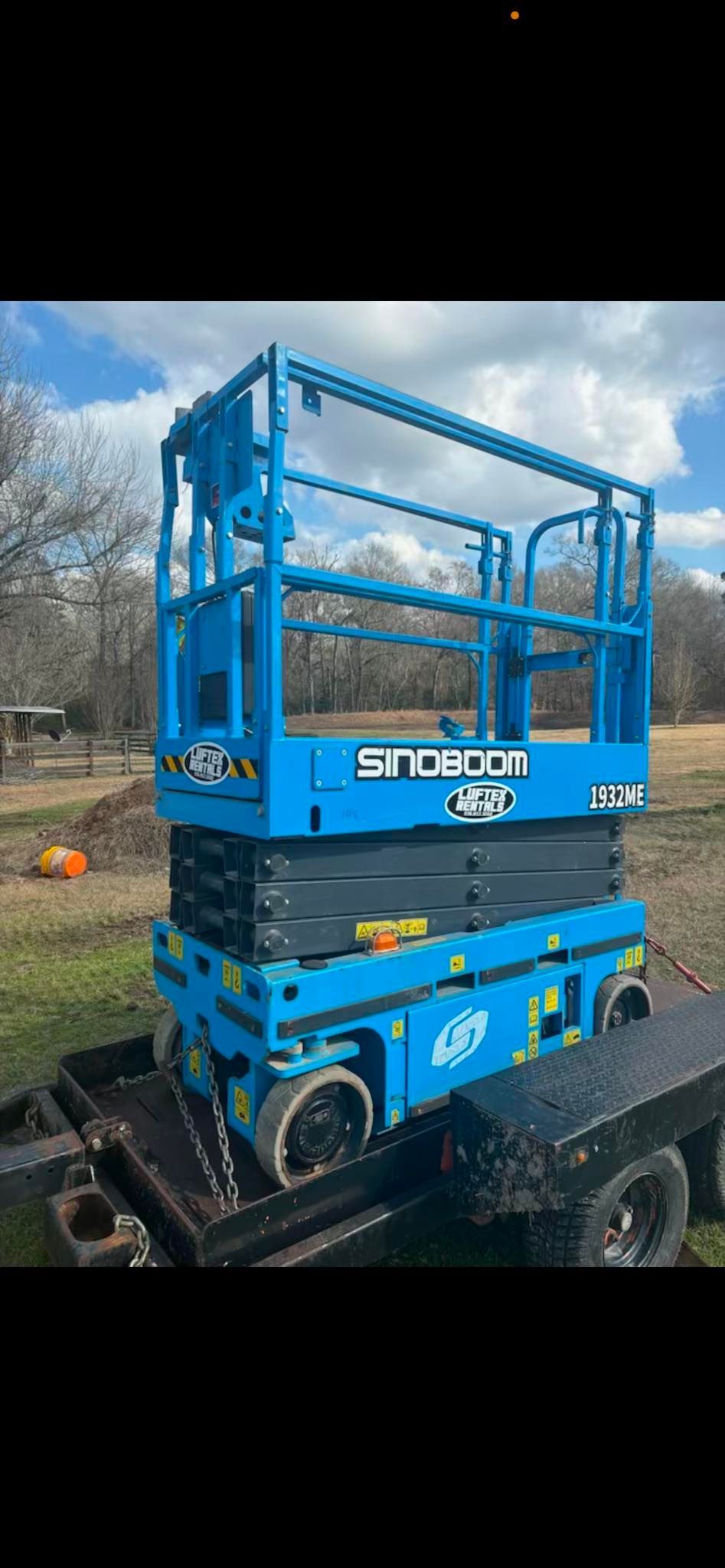 A bright blue Sinoboom scissor lift secured on a flatbed trailer in an outdoor field.