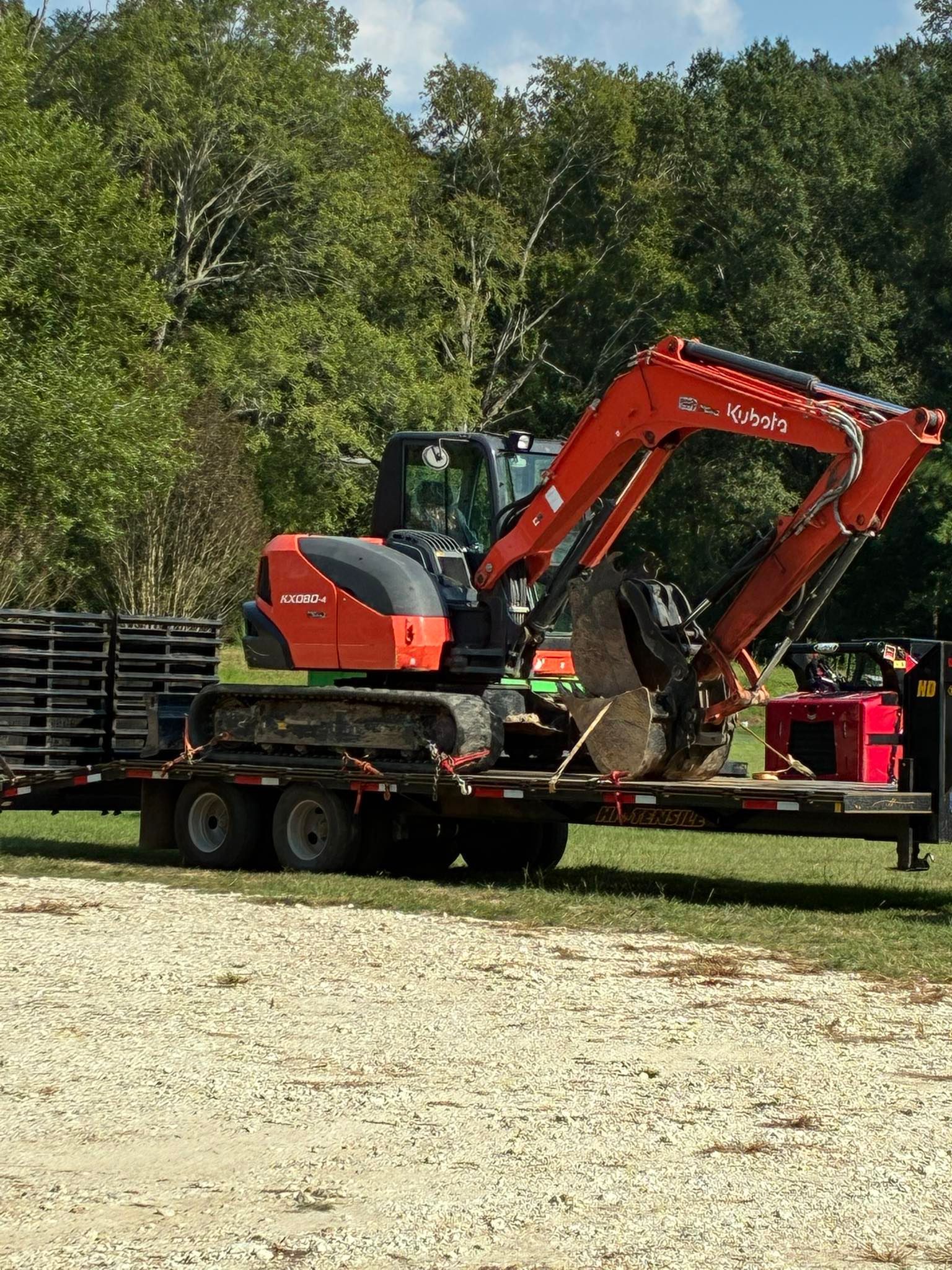 An orange Kubota excavator sits on a flatbed trailer in a grassy, wooded area.