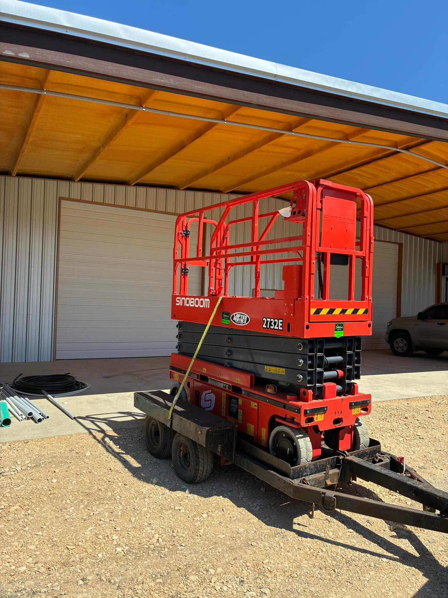 A red scissor lift is loaded onto a flatbed utility trailer parked in a gravel lot in front of a metal workshop building.