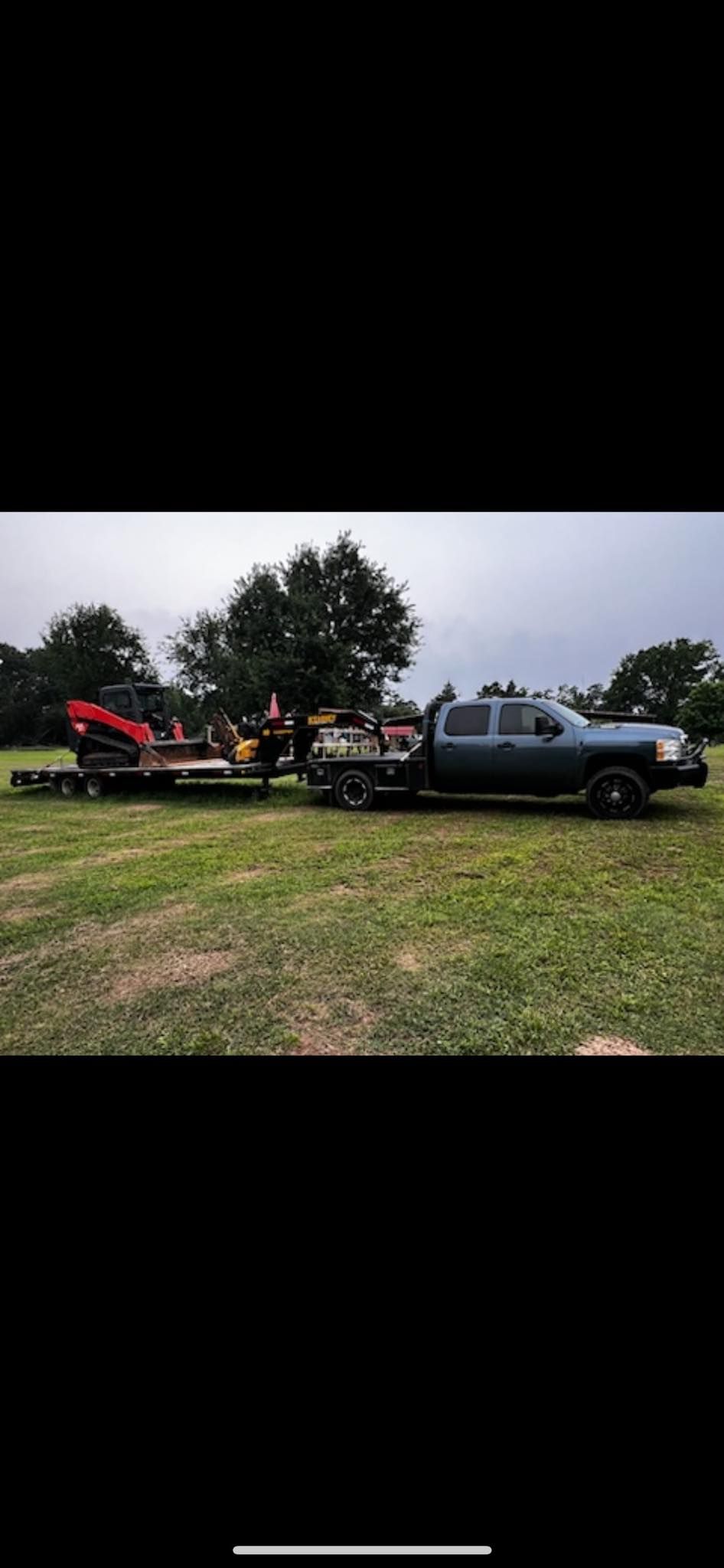 A dark-colored pickup truck with a flatbed trailer parked in a field, hauling a small red piece of construction equipment.