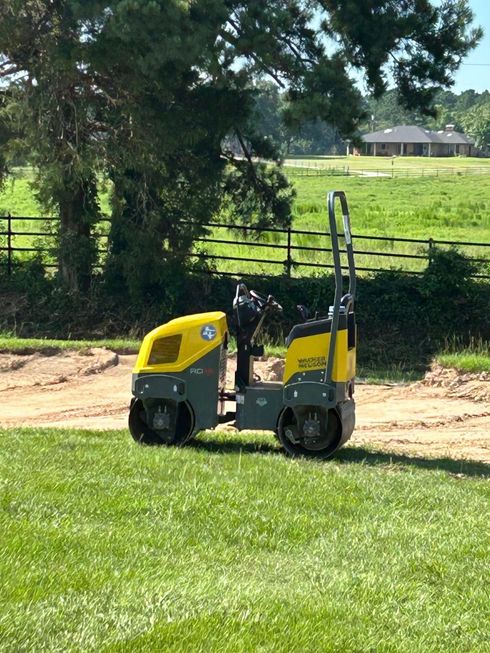 A yellow and grey tandem vibratory roller sits on a grass lawn near a dirt construction area with a rural backdrop.