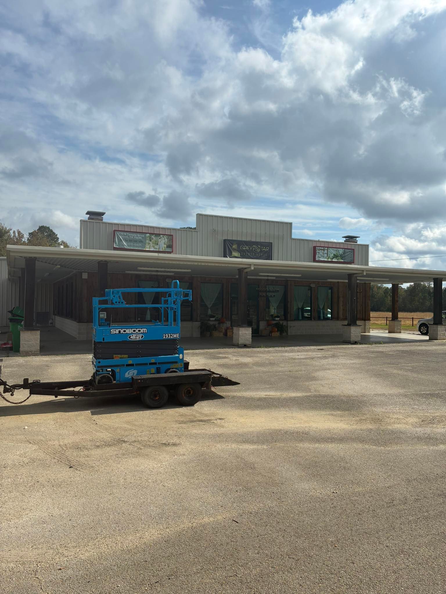 A blue scissor lift on a trailer parked in front of a rustic, single-story commercial building under a cloudy sky.