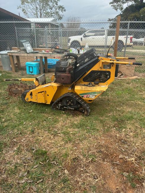 A yellow walk-behind stump grinder with black tracks sits on a grass lawn in front of a chain-link fence.