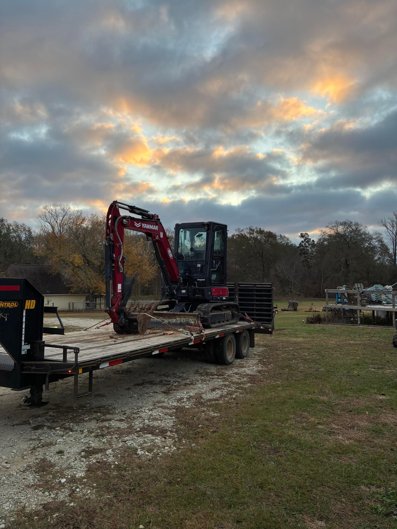 A red and black excavator sits loaded on a flatbed trailer in a grassy, open area under a cloudy sunrise.