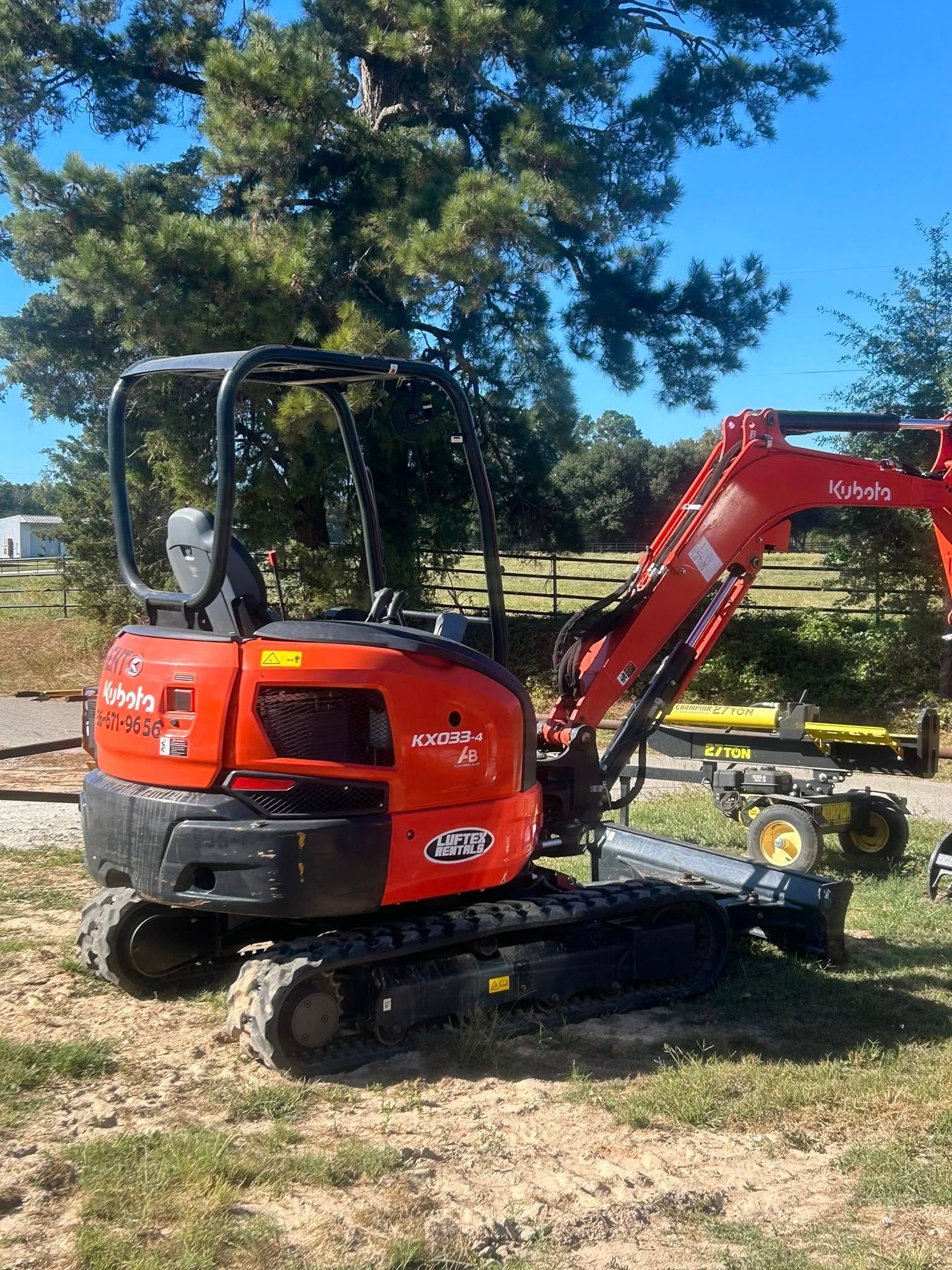A bright orange Kubota mini excavator parked on grass and dirt in front of a tree on a sunny day.