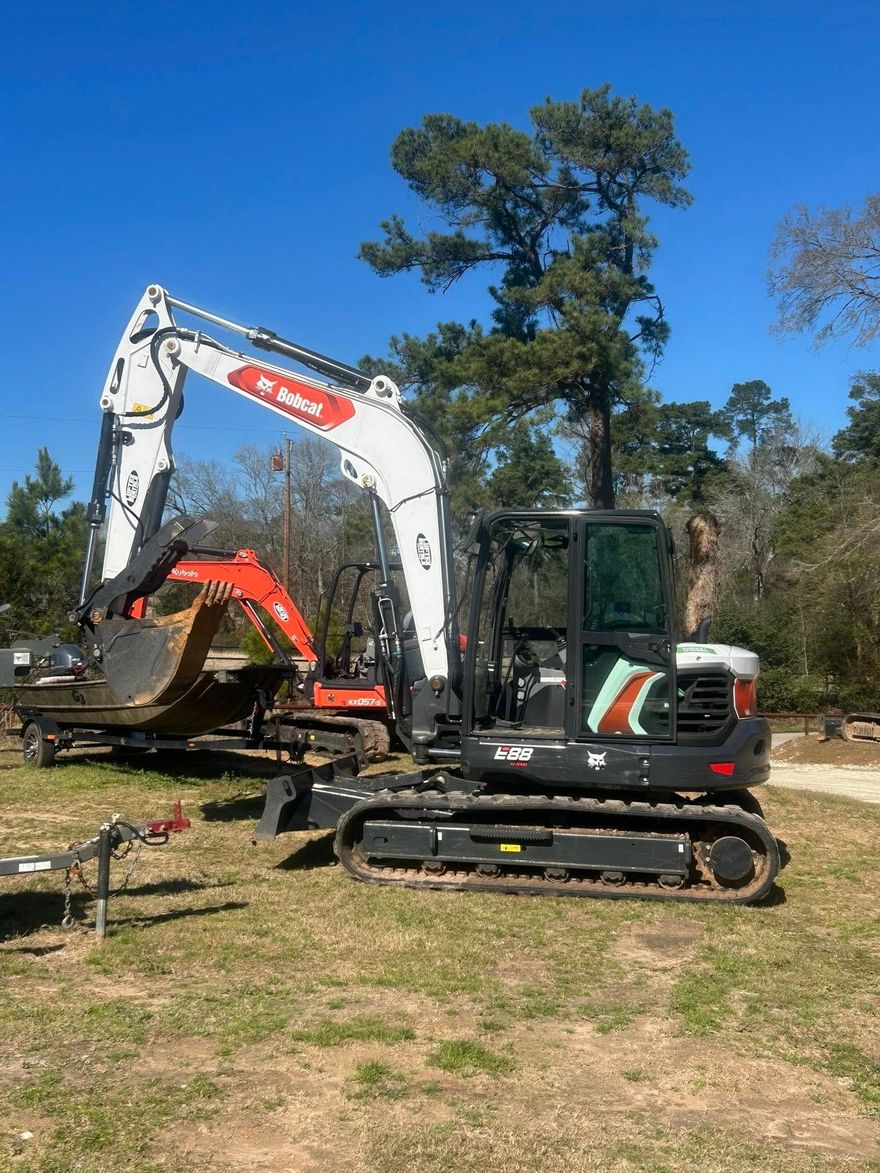 A white and black excavator parked on a grassy field, with a smaller orange piece of equipment on a trailer behind it.