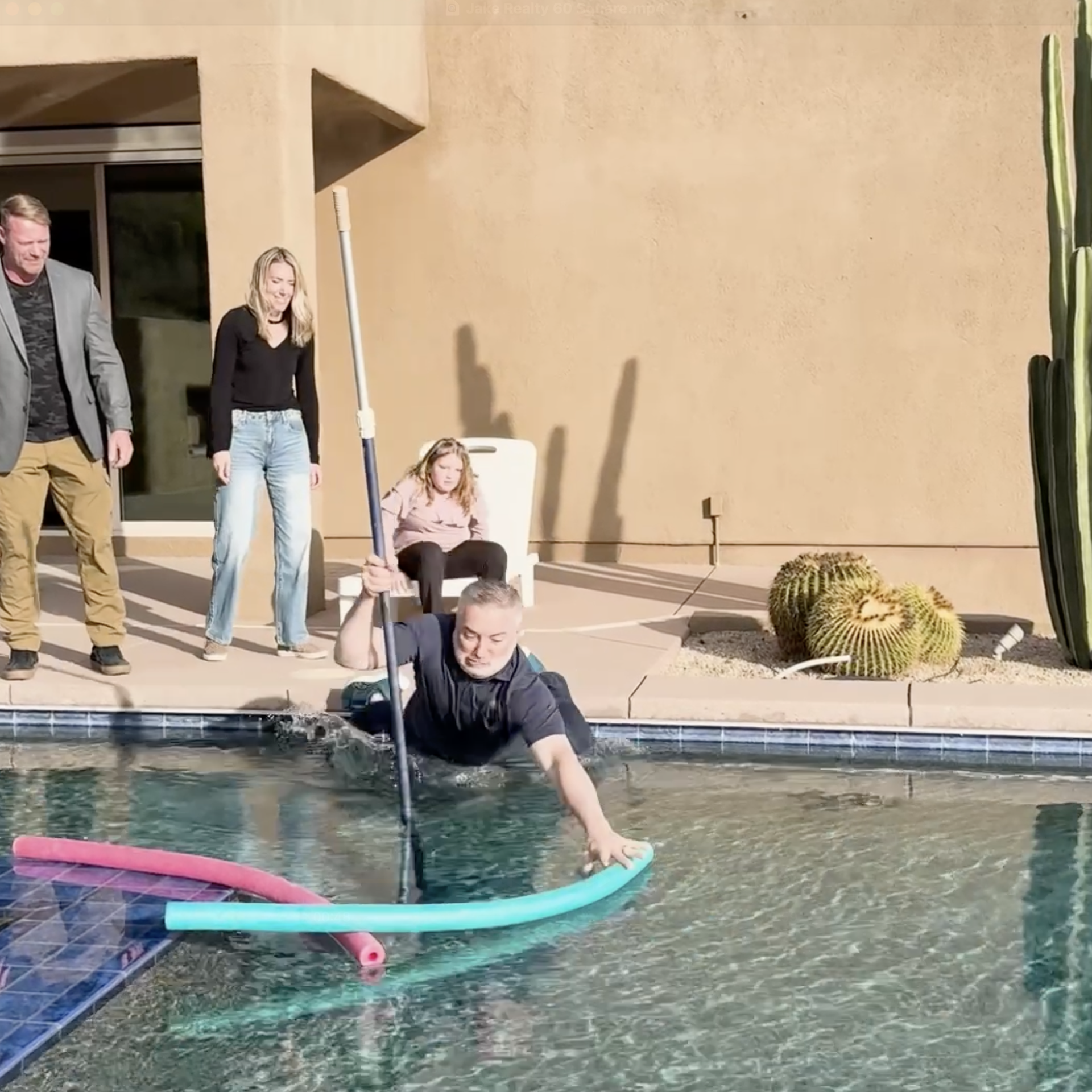 Back yard, man falling in a pool holding a pool cleaning tool and reaching for a pool noodle. 