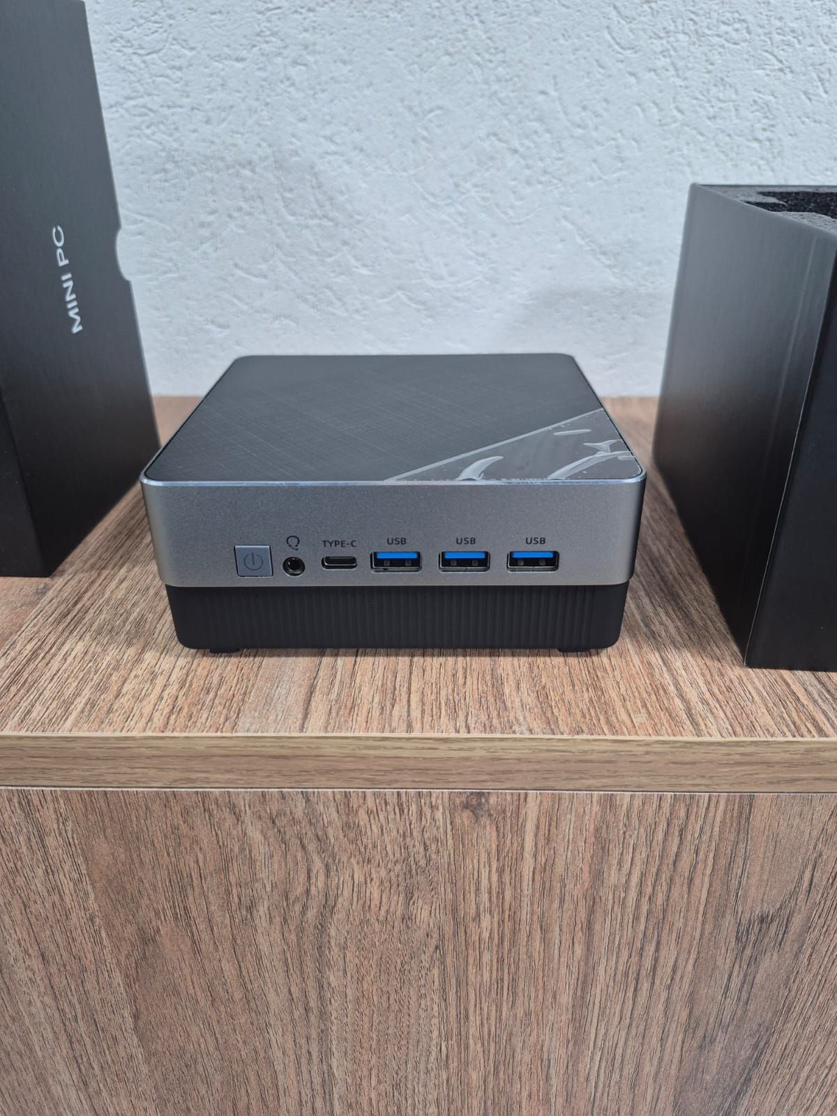 Mini PC negro de pequeño tamaño sobre una repisa de madera, visto desde el frente con los puertos visibles.