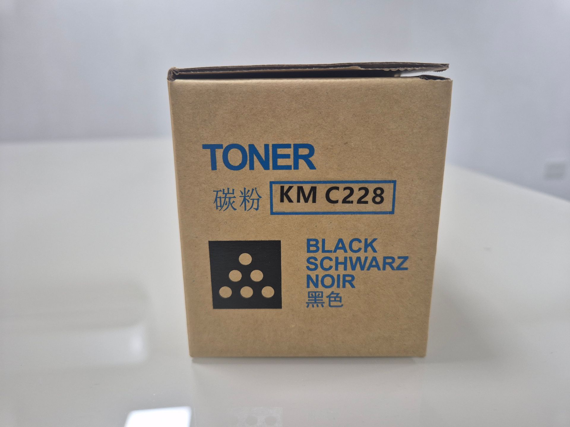 Caja de cartucho de tóner marrón con la etiqueta “TONER KM C228” y “BLACK/SCHWARZ” sobre una superficie blanca.