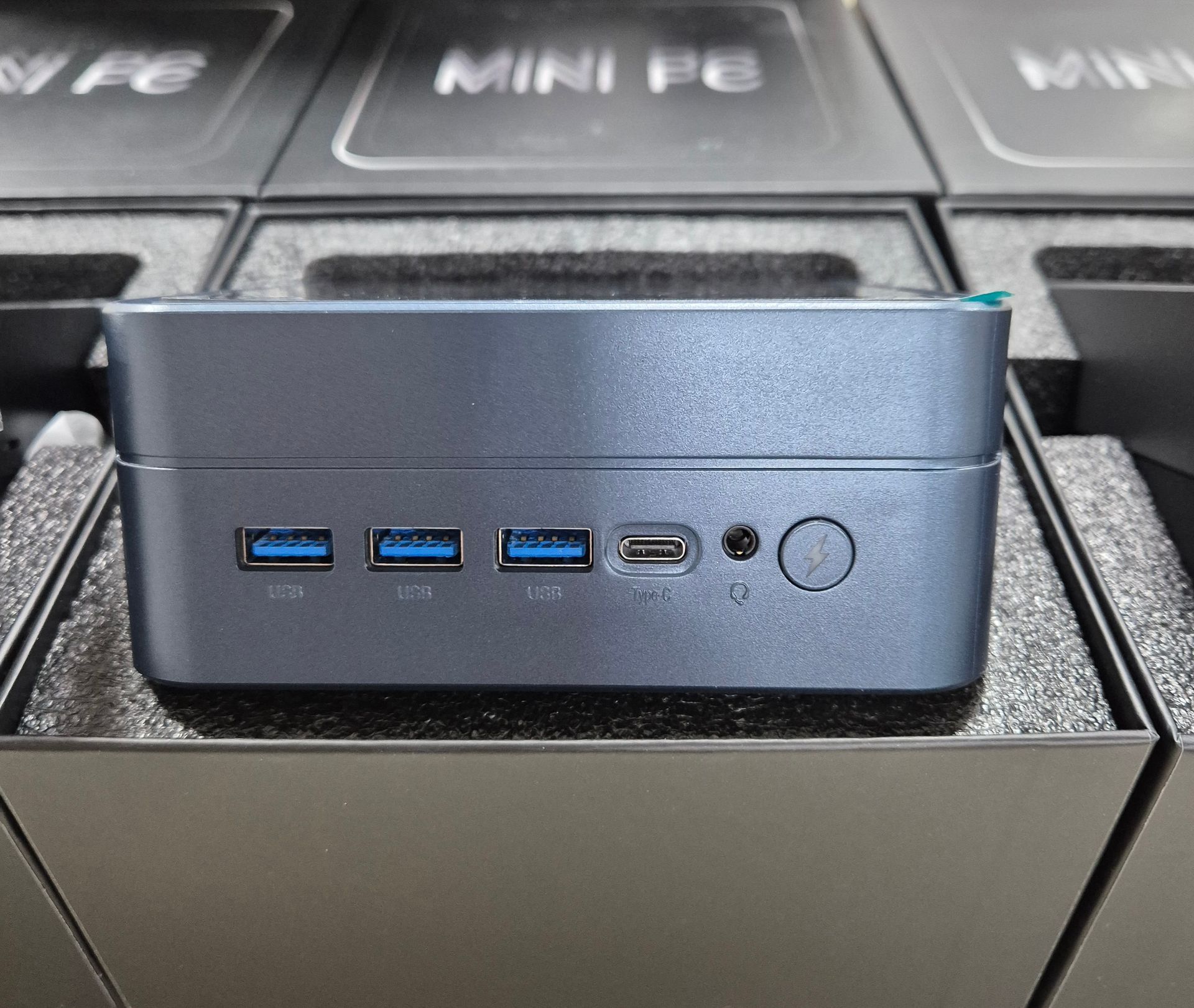 Mini PC pequeño de color gris con puertos USB azules y botón de encendido, dentro de una espuma de embalaje.
