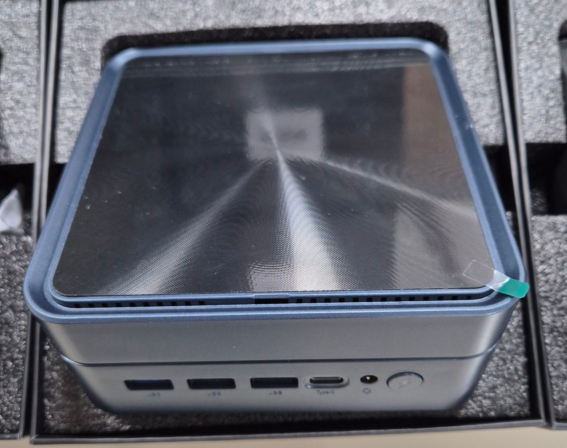 Mini PC pequeño de color gris con puertos USB azules y botón de encendido, dentro de una espuma de embalaje.