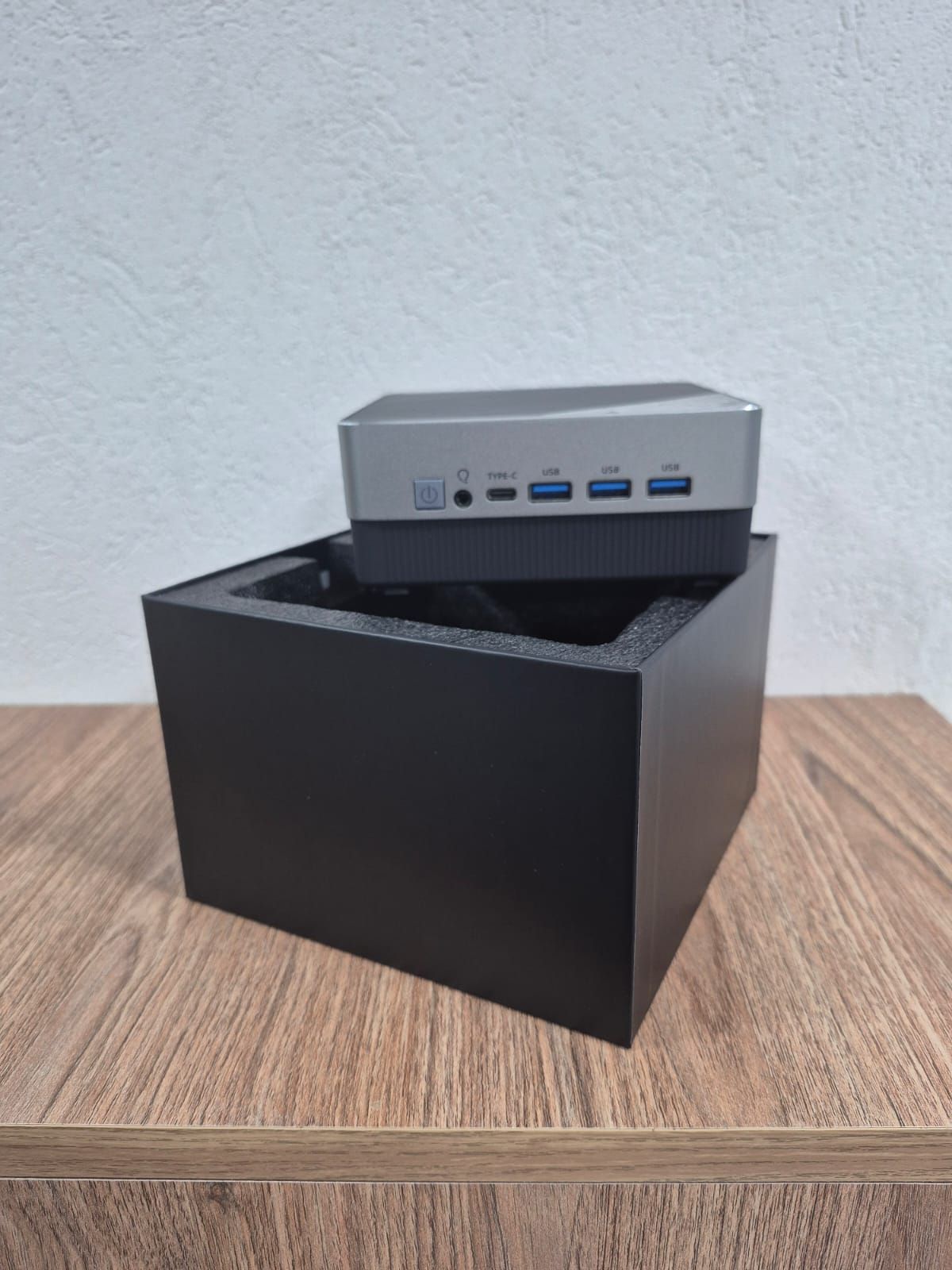 Mini PC pequeño de color plateado descansa sobre una caja negra encima de una mesa de madera, contra una pared clara.