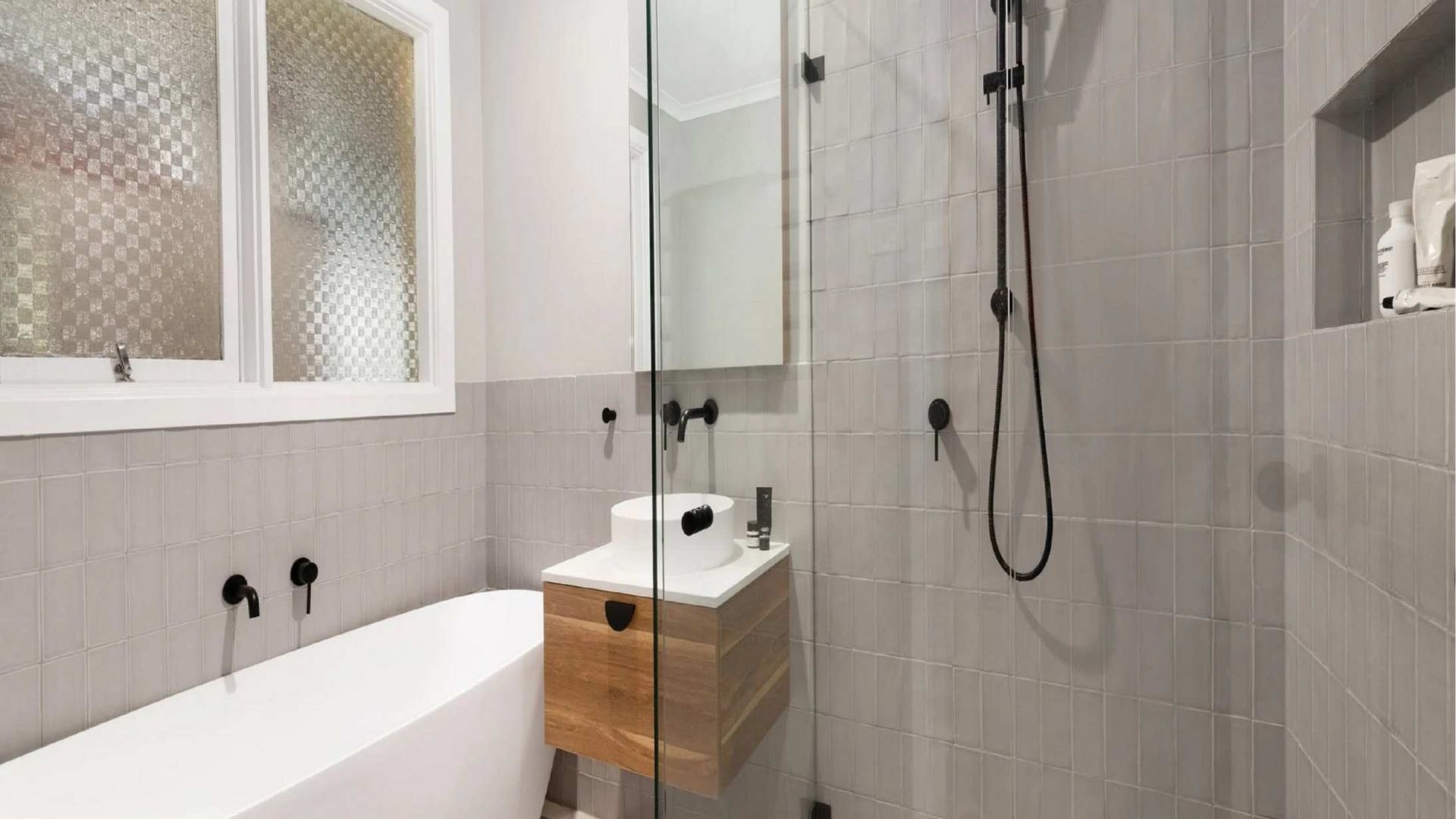 Modern bathroom with gray tiled walls, a white soaking tub, a wood-toned floating vanity, and a glass-enclosed shower.