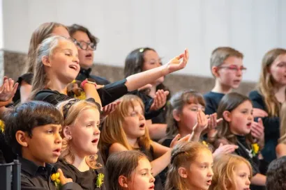 A choir of children singing in unison, with some raising their hands while performing in a bright indoor space.